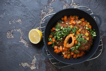 Cast-iron pan with ossobuco made of cross cut veal shank, view from above on a brown stone background, copyspace