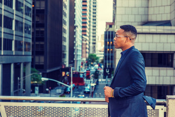 African American Businessman working in New York. Wearing blue jacket, glasses, a black man standing by railing on balcony, facing street, sad, thinking, lost in thought. Filtered effect..