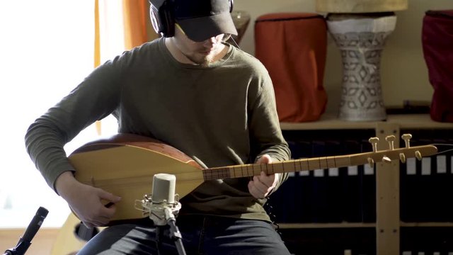 Medium tight shot of a young man wearing headphones plays a baglama saz with microphones in front of instrument and doumbek and oud in background.