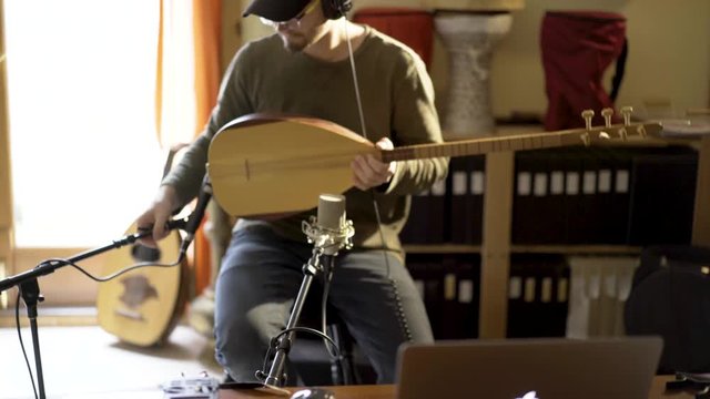 While holding a saz a young man adjusts computer and microphones and sits down to start recording instrument.