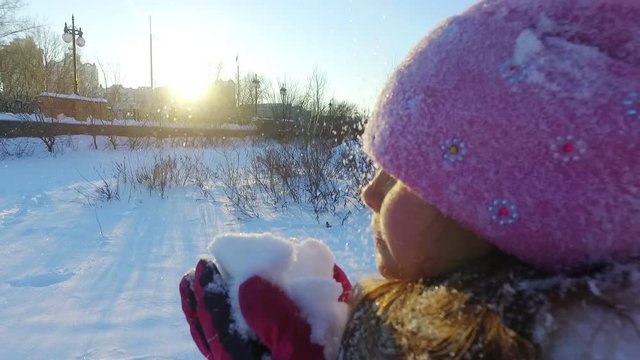 Close up of little girl blowing snow from hands. Kid playing with snow in slow motion. Winter wonderland. Portrait of winter girl holding snow