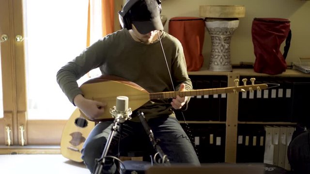Young man wearing headphones playing baglama saz with microphones in front of instrument and oud and darbuka drums in background in a room with a lot of light.