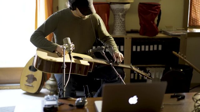 Wide camera slide to right as young man wearing headphones and playing slide guitar with microphones and computer in home recording studio.