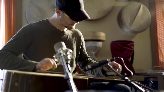 Young man with ball cap playing music on a slide guitar.  Doumbek and frame drums in the background.