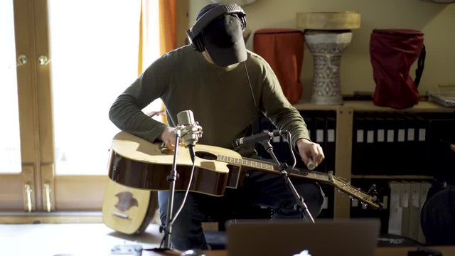 Wide camera slide to right as young man wearing headphones and playing slide guitar with microphones and computer in home recording studio.