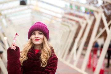 Beautiful blonde girl in red knitted hat posing with candy cane at the Christmas fair