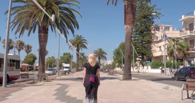 ALGHERO, SARDINIA, ITALY  &ndash; JULY 2016 : Video shot of beautiful blonde girl walking on main street on a sunny day with palm trees and traffic in view