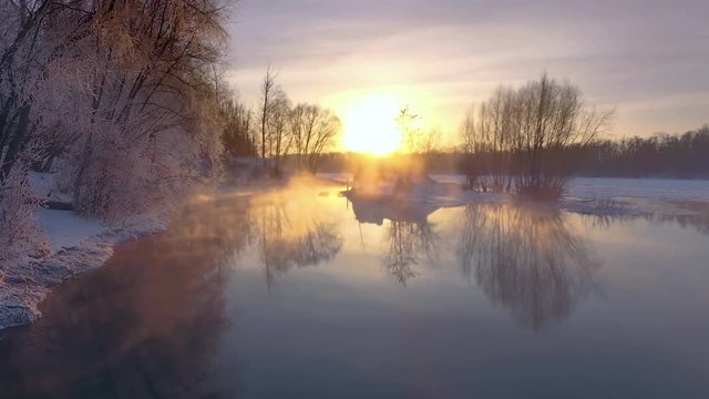 AERIAL: Peaceful winter landscape during sunrise. Snowy forest, frozen river, steam above the water. Beautiful reflections on water surface, colorful winter tale.