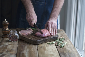 a man in a kitchen apron is cutting meat with a knife on a board and a wooden table, rosemary and spices
