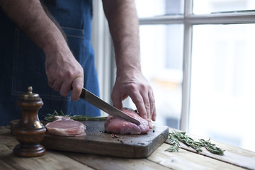 raw pork meat on a wooden board, spices and rosemary, the hands of a male cook keeps a knife and slice of meat. near the window