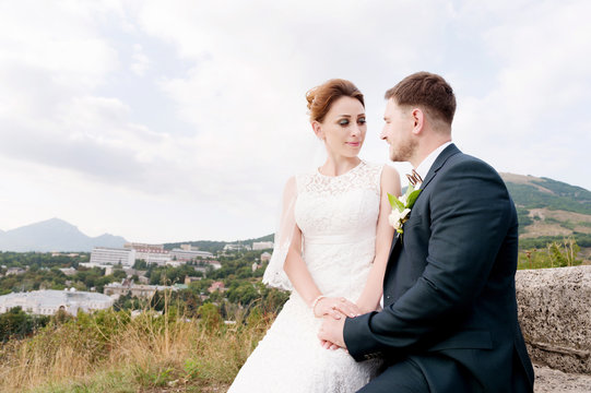 A loving couple of newlyweds sits on the background of the cityscape of a small resort town in the Caucasus. Newly married gently caress each other