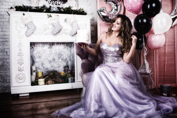 a chic young woman with a charming smile in a beautiful dress with bare legs sitting on a chair in front of a fireplace and a Christmas tree. beautiful interior