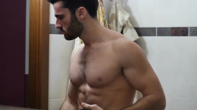 Attractive muscular young man dressing, putting on black t-shirt in bathroom, hiding naked ripped torso and muscles on chest, abs, arms