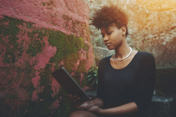 Portrait of young dazzling African American girl sitting on the staircase near red wall with digital pad; serious curly black lady with tablet pc outdoors on the concrete stairway in dark settings