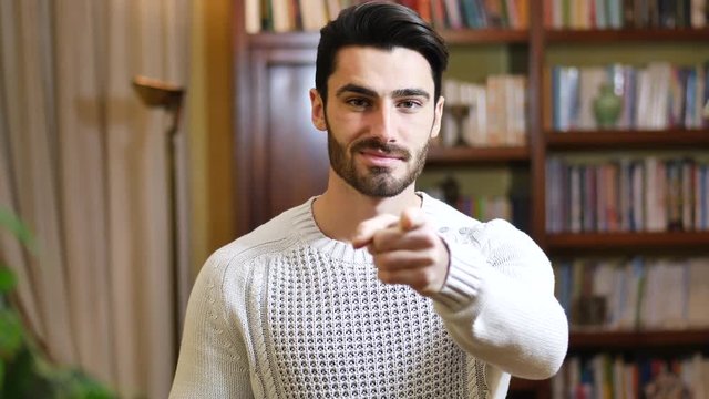 Handsome young man pointing finger at you, smiling. Indoors shot inside a house