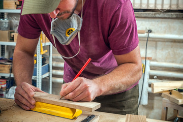 A young strong man builder carpenter works with a wooden bar for making furniture, measures and cuts in the workshop, in the background of many tools and working conditions