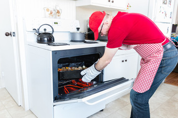 horizontal image of a man wearing a gingham red apron and a red hat and shirt bending over the oven...