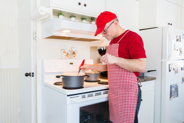 horizontal image of a man wearing a gingham apron and red shirt and hat holding a glass of wine and...