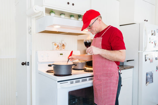 Horizontal Image Of A Man Wearing A Gingham Apron And Red Shirt And Hat Holding A Glass Of Wine And Stirring The Pot On The Stove Preparing A Meal