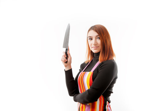 A young red-haired woman in a black turtleneck and a kitchen apron posing and holding a large kitchen knife on a white isolated background