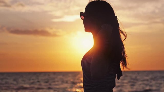 Silhouette of seductive girl with long hair wearing sunglasses enjoying the sunset on the beach in slow motion in windy weather by the sea. 1920x1080