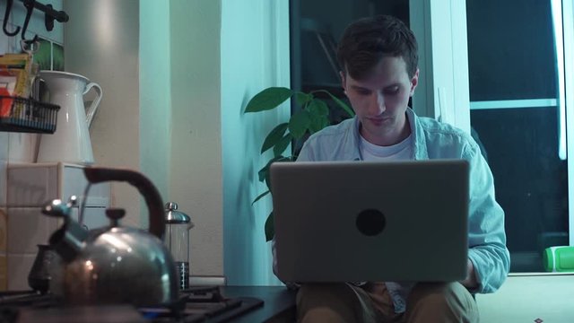 Young man using computer sitting at kitchen while kettle boiling on stove