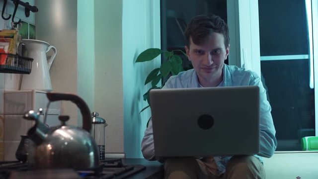 Young guy using computer sitting at kitchen while kettle boiling on stove