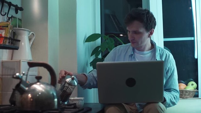 Young guy typing on computer sitting at kitchen while kettle boiling on stove