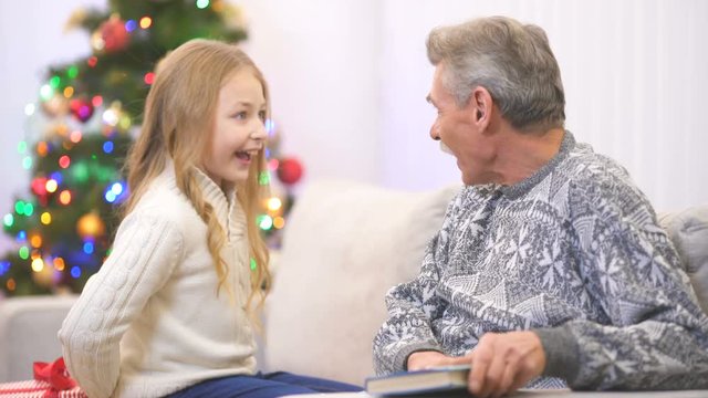 The grandfather read a book and take a gift from a girl near the christmas tree