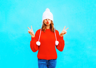 Obraz premium Pretty young woman with red lips does an air kiss in a knitted hat, sweater on blue background