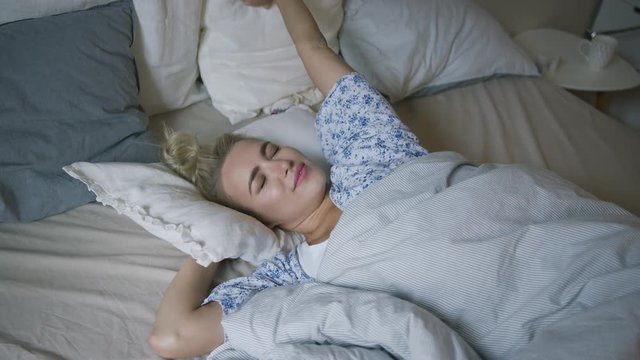 Attractive young woman in shirt lying on comfortable bed under soft blanket and stretching arms in morning. 