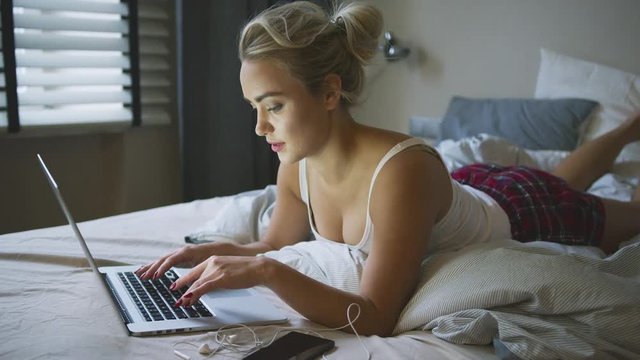 Attractive young woman in tank top and shorts lying on bed and browsing laptop in morning. 