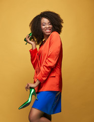 Bright portrait of young african girl with afro hairstyle. Smiling girl wearing red orange jacket and violet latex skirt holds green heels in her hands and posing on yellow background. Studio shot.