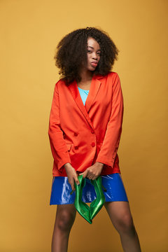 Bright portrait of young african girl with afro hairstyle. Girl wearing red orange jacket and violet latex skirt holds green heels in her hands and posing on yellow background. Studio shot.