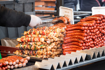 Chef with metal pincers holds 2 sausages and removes them from grill. There is one hill from sasages and and another one from shish kebab skewer.