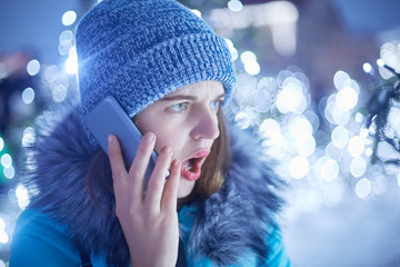 Surprised woman dressed in winter clothes, opens mouth in bewilderrment, speaks with her boyfriend via smart phone, doesn`t expect to hear shocking news, has walk outdoor. City decorated with lights