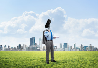 Camera headed man standing on green grass against modern cityscape