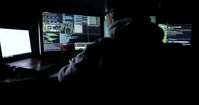 Male hacker in a hood works on a computer with maps and data on display screens in a dark office room