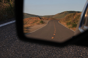 Looking into a rearview mirror at a landscape image of a deserted, divided highway heading towards the Northern California foothills near Petrolia.