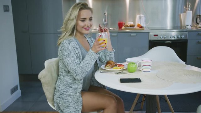 Cheerful young woman sitting at round kitchen table and drinking orange juice while enjoying healthy breakfast.
