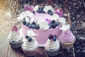 Purple beautiful cake decorated with berries, blackberries and blueberries on top with cupcakes on...