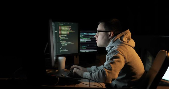 Hacker in hood cracking code using laptop and computers from his dark hacker room