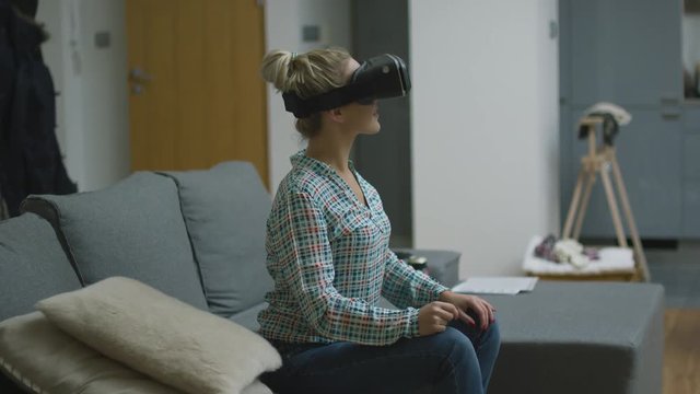 Beautiful young woman in checkered shirt sitting on sofa and wearing VR headset with astonished face expression.