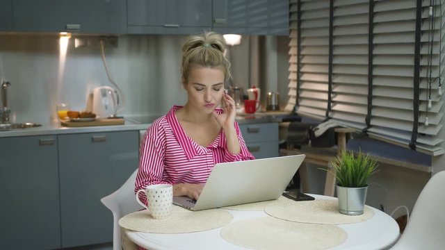 Lovely young woman in striped pink bathrobe sitting at round kitchen table and using laptop in morning.