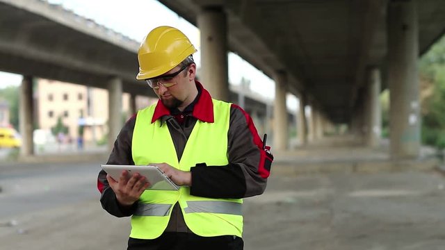 Workman with tablet computer under overpass. Manager of the repair works at construction site under overhead road, inputs data in his computer
