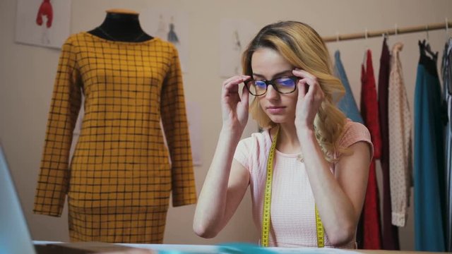 Portrait of the blonde female designer taking on the glasses and starting to work at the laptop computer on the new clothes collection. Mannequin in the yellow dress on the background. Indoors