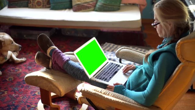 Steadicam Closeup Shot Of Woman Working On Laptop Computer With Green Screen And Dog At Her Feet Curled Up On Rug.