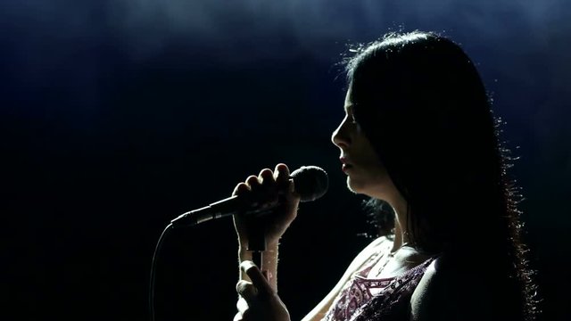 Portrait of a beautiful singer with long hair on a dark smoky stage in the spotlight. Female singer on the stage holding a microphone.