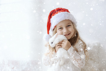 cute little girl in a white dress with a white little rabbit in a santa claus hat