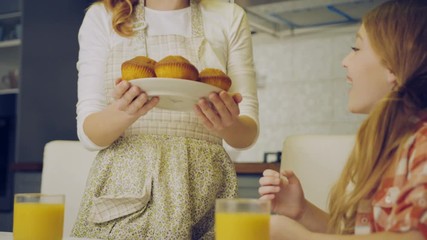 Beautiful mother in the apron bringing and putting on the kitchen table muffins on the plate and her lovely daughter hugging her. Mom kissing a girl on the forehead. Indoors - Powered by Adobe